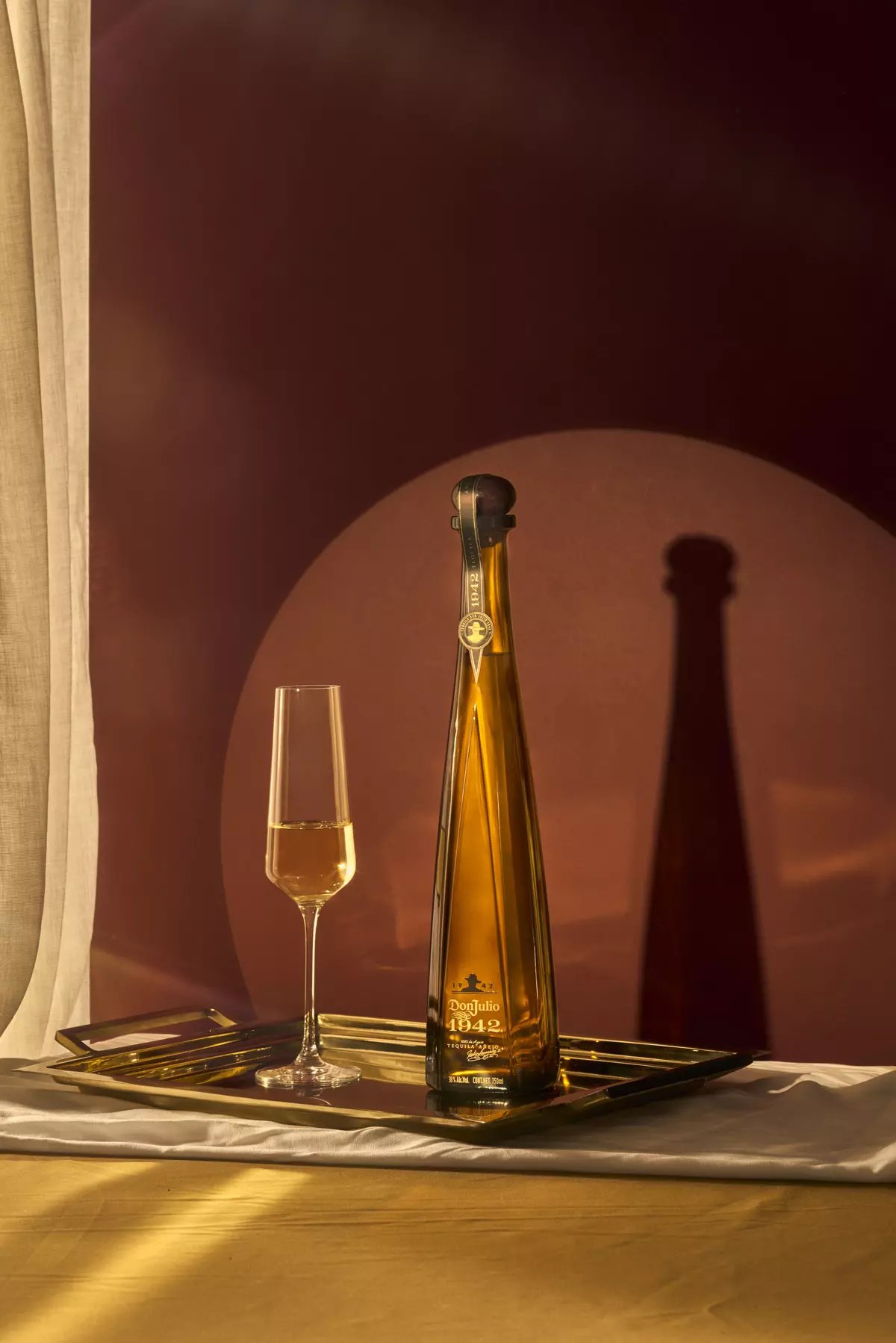 A tall, slender bottle of golden tequila and a filled flute glass are displayed on a metallic tray with a dramatic shadow against a burgundy background.
