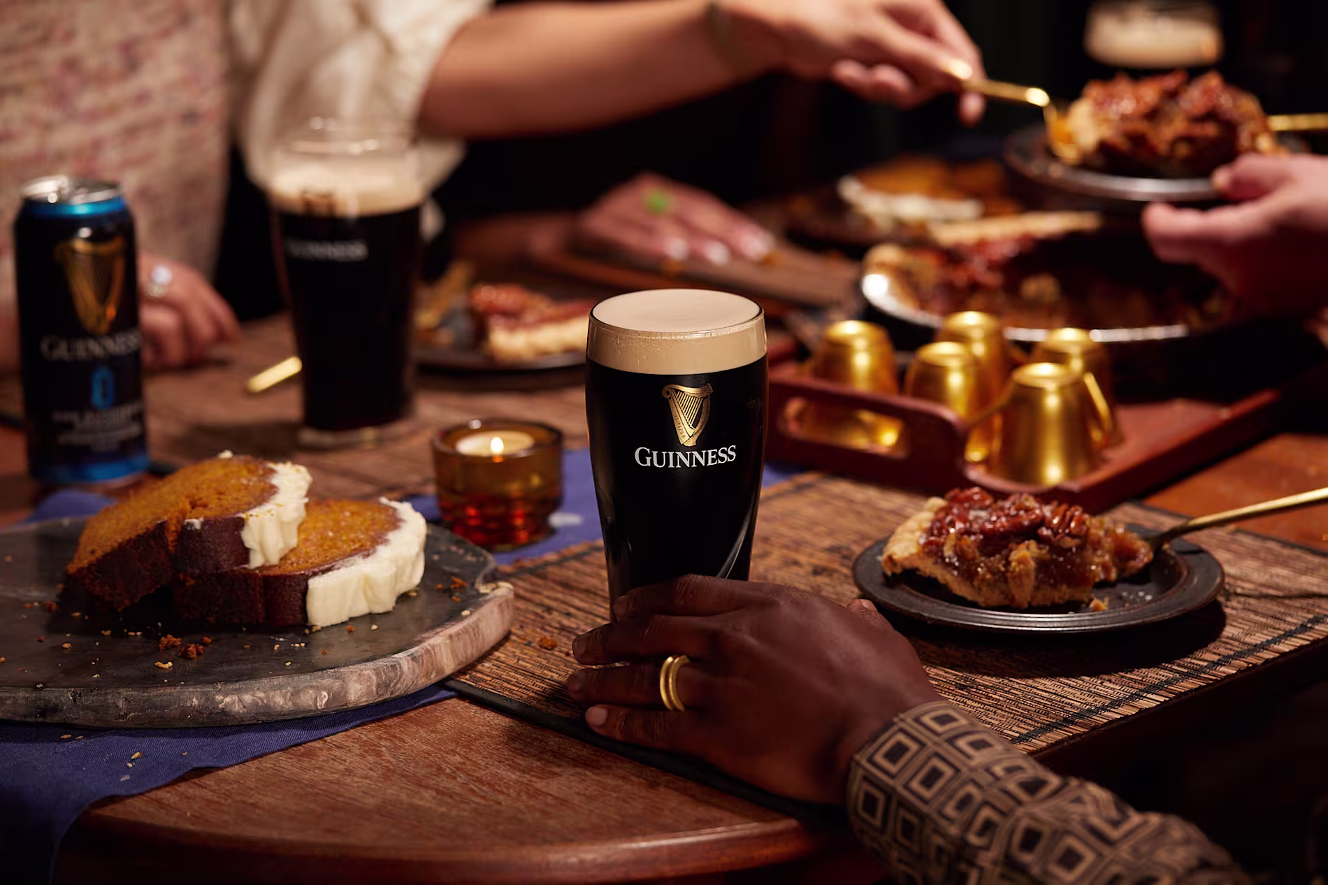 Pint of Guinness stout on a wooden table with slices of cake and pecan pie, surrounded by people enjoying dessert