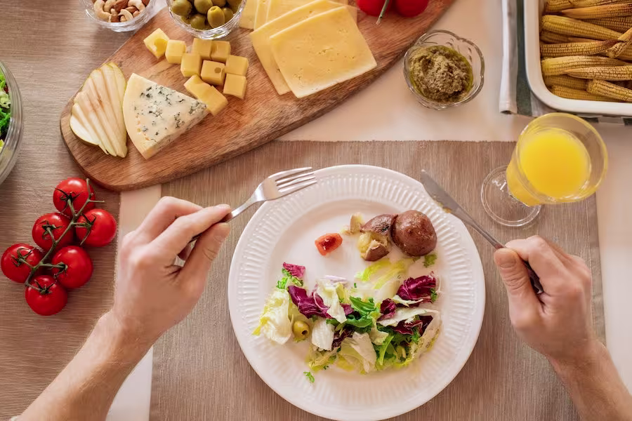Person holding fork and knife over a plate of salad with greens, olives, lettuce, and a potato; cheese, bread, juice, tomatoes, and other sides are on the table.