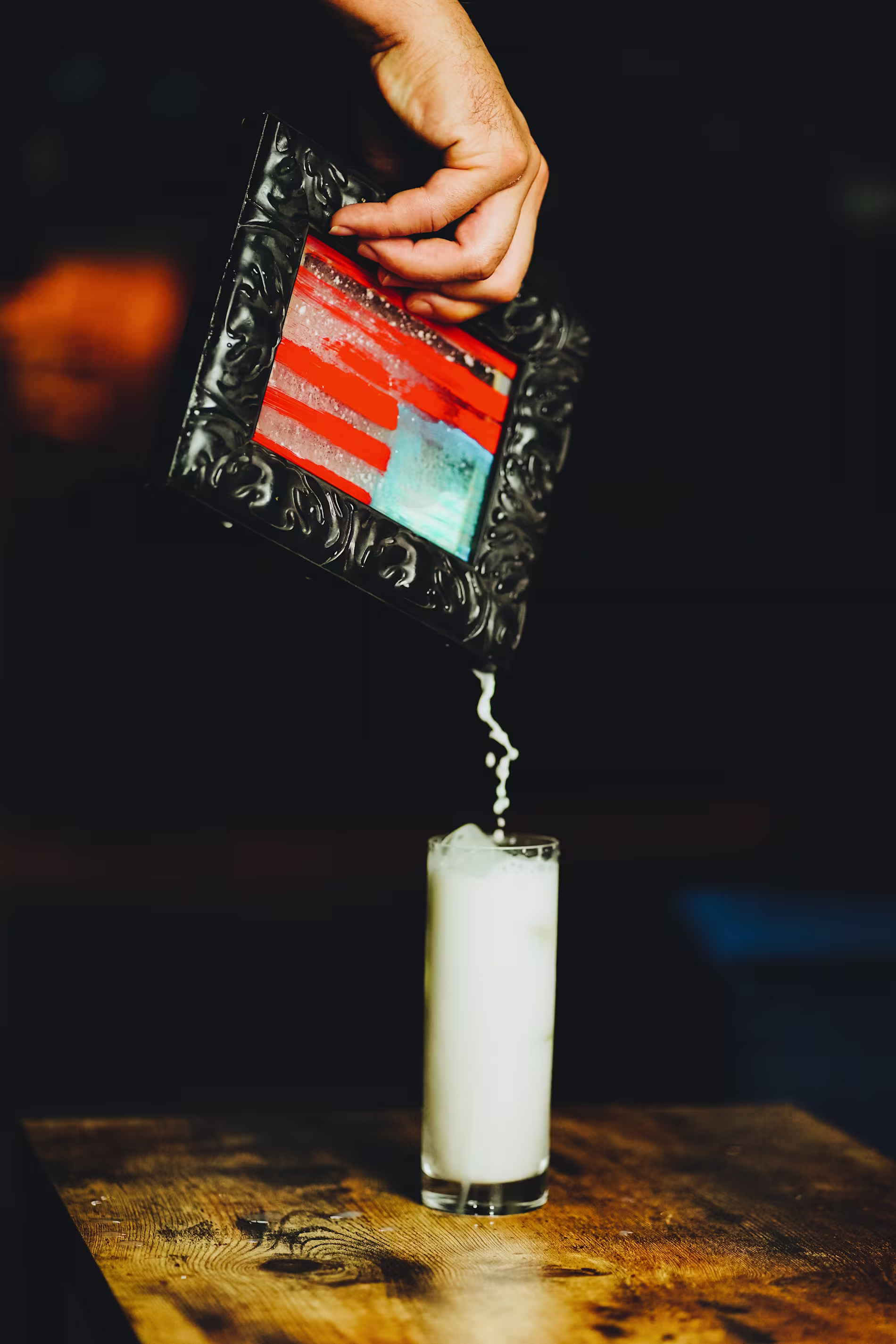  Una mano sosteniendo un pequeño marco negro ornamentado, desde el cual se vierte un líquido en un vaso de cocktail. El marco tiene rayas rojas y blancas, que recuerdan al patrón de la bandera estadounidense. 