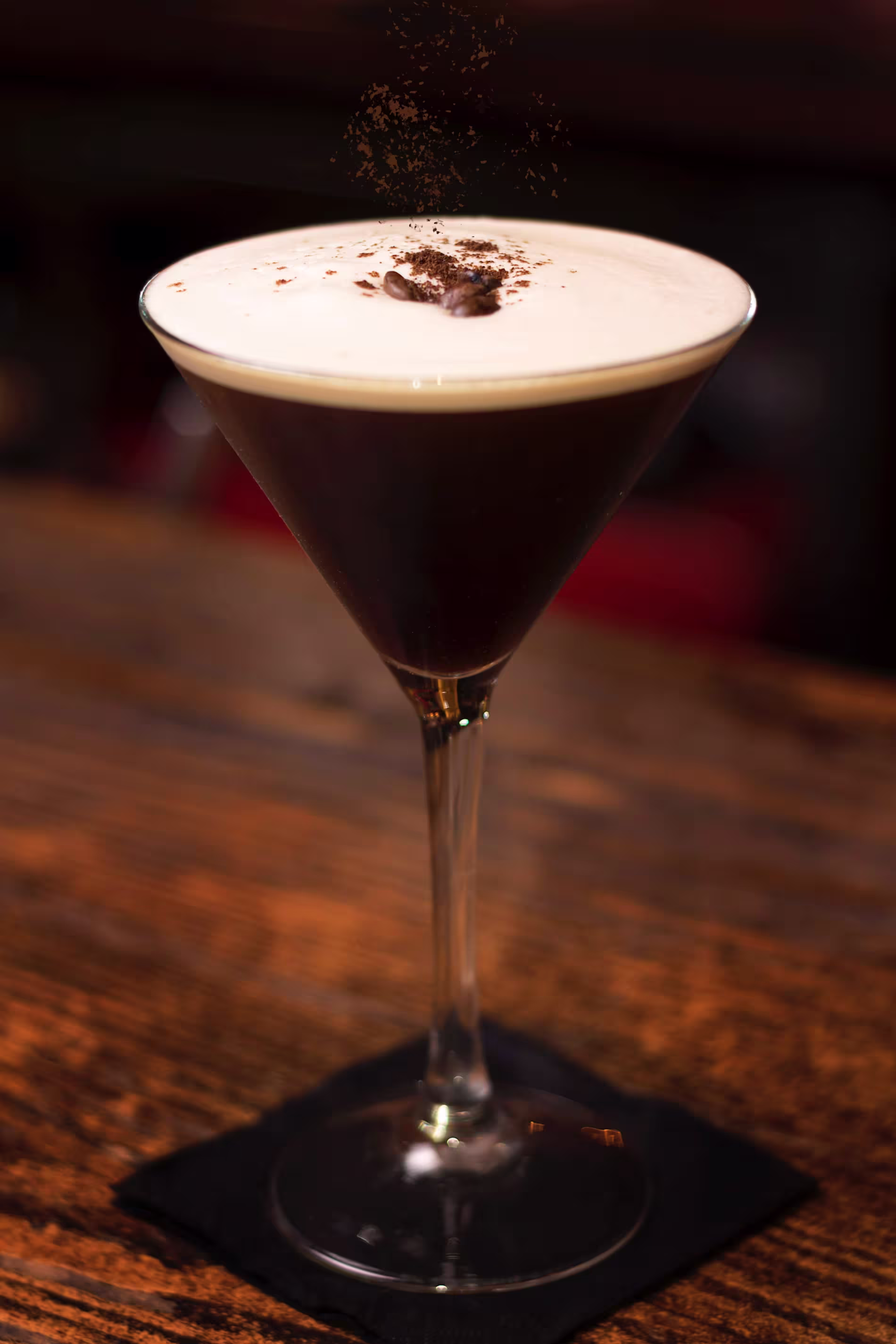A glass of espresso martini with a foamy top, garnished with coffee beans, sits on a wooden surface.