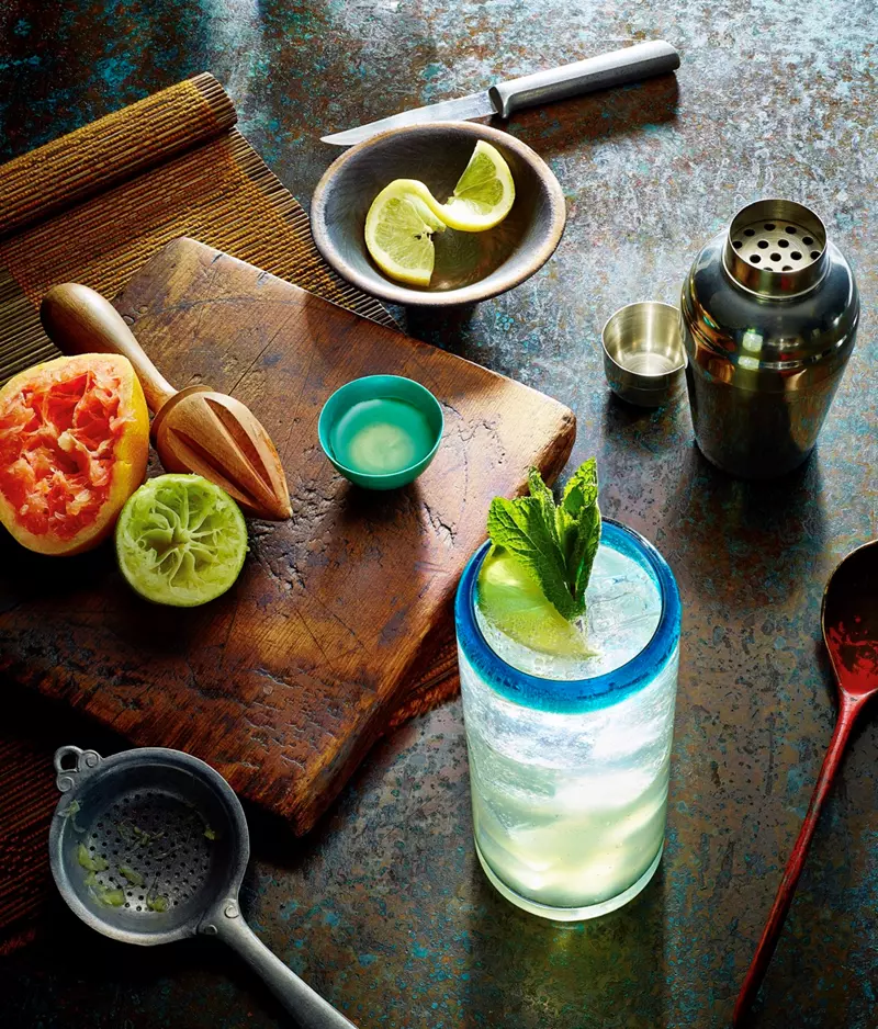 A glass of citrus cocktail with mint sits on a table, surrounded by a cutting board with halved citrus fruits, a knife, a shaker, strainer, and a plate with lime wedges.