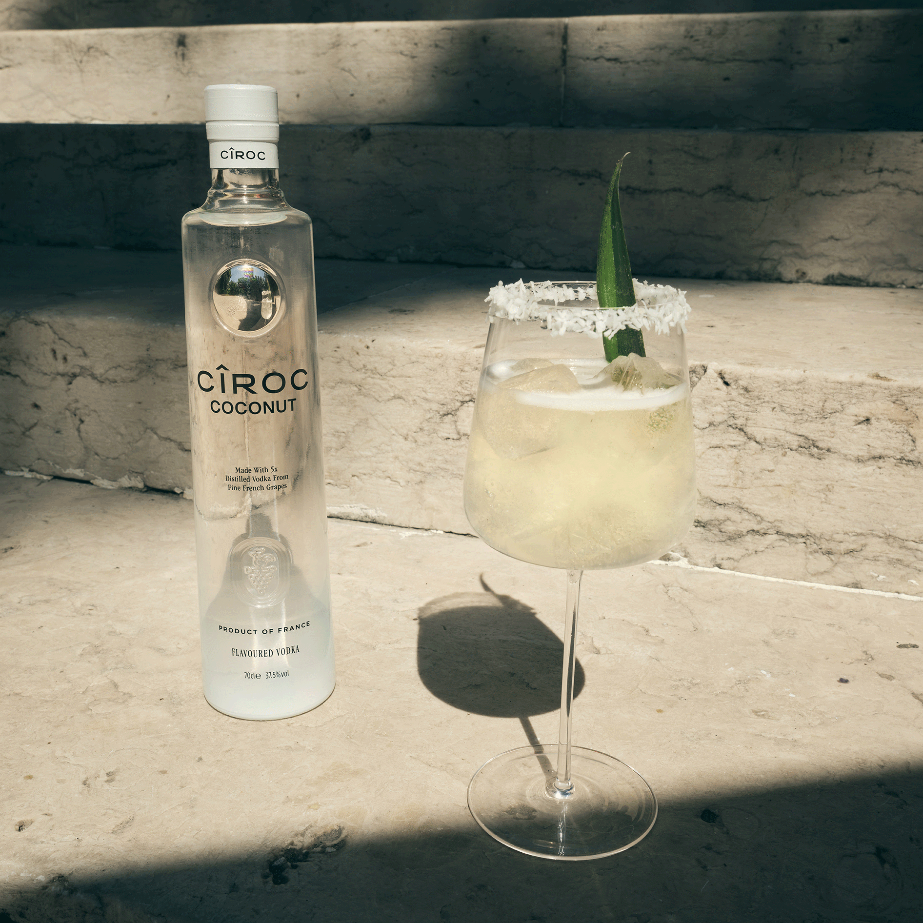 A bottle of Ciroc Coconut vodka stands next to a cocktail in a wine glass with a salted rim and pineapple leaf garnish, placed on stone steps in sunlight.