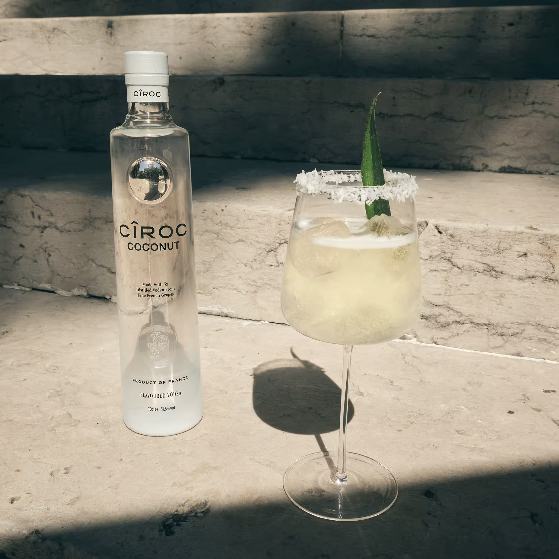A bottle of Ciroc Coconut vodka stands next to a cocktail in a wine glass with a salted rim and pineapple leaf garnish, placed on stone steps in sunlight.