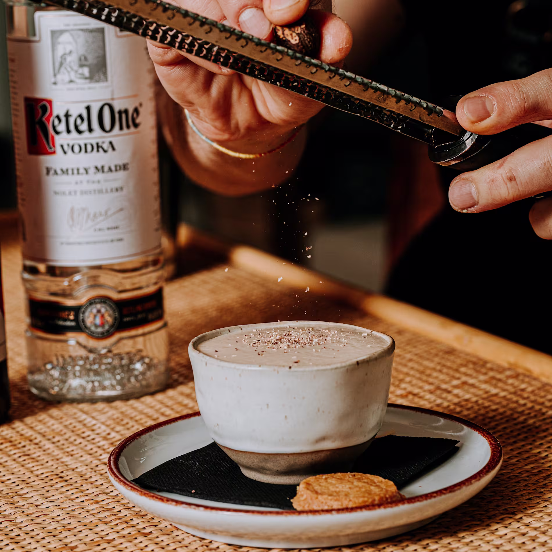 Manos rallando nuez moscada sobre una bebida cremosa servida en taza de cerámica. Al fondo, botella de Ketel One Vodka sobre una mesa de mimbre.