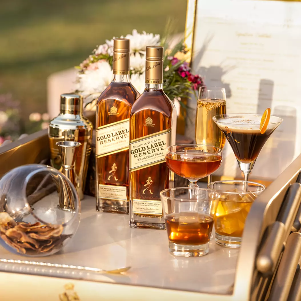 Two bottles of Johnnie Walker Gold Label Reserve whisky on a bar cart with cocktails, a shaker, dried citrus, and flowers in the background.