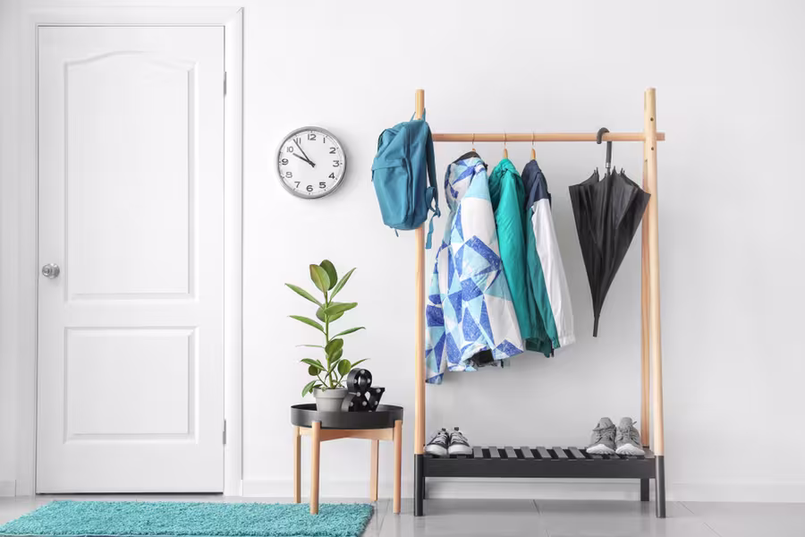 A coat rack with jackets, shirts, a backpack, and umbrellas stands beside a small table with a plant and decor; shoes are below the rack and a wall clock hangs nearby.