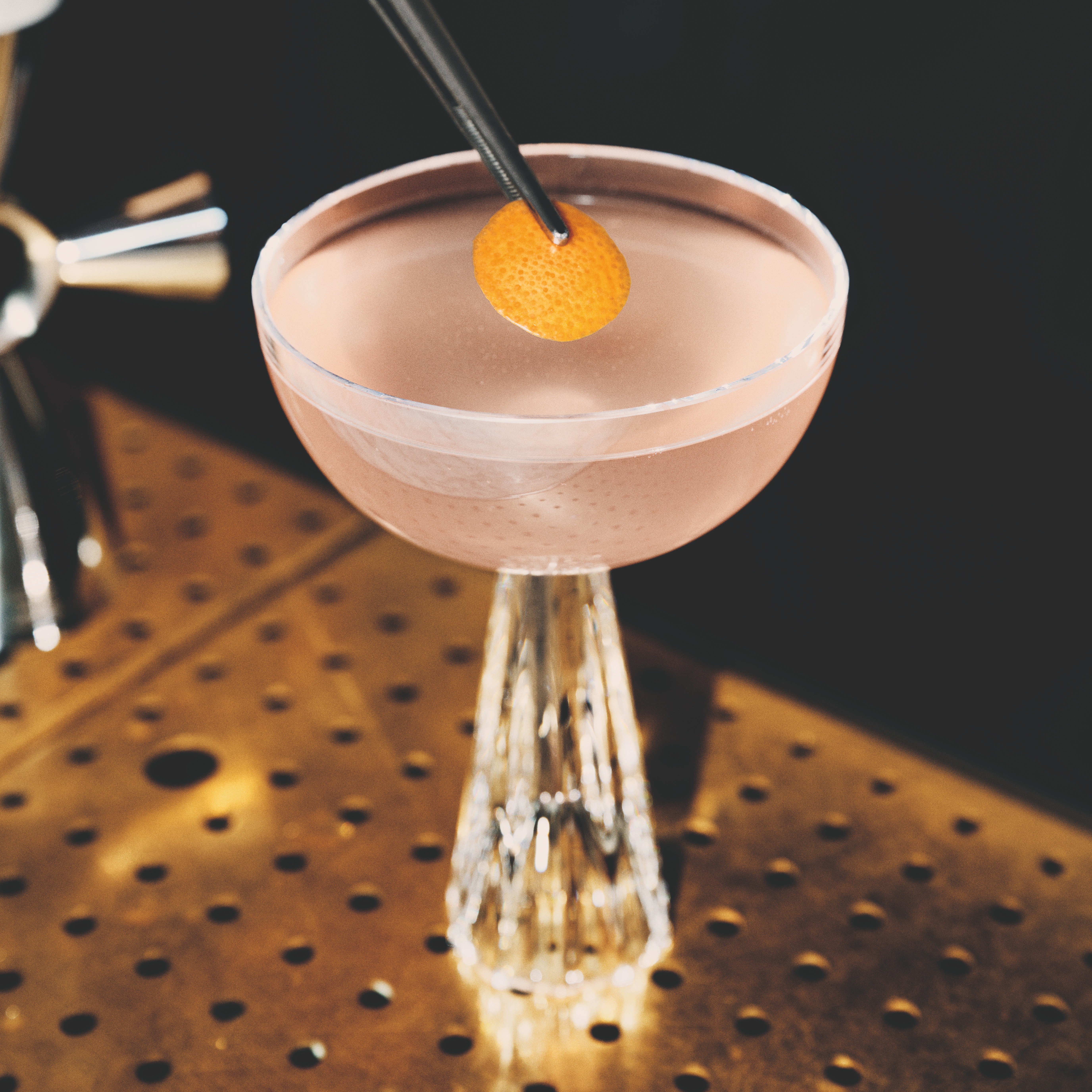 A small orange garnish is placed on a pastel pink Tiny Ten cocktail in a fancy coupe glass.