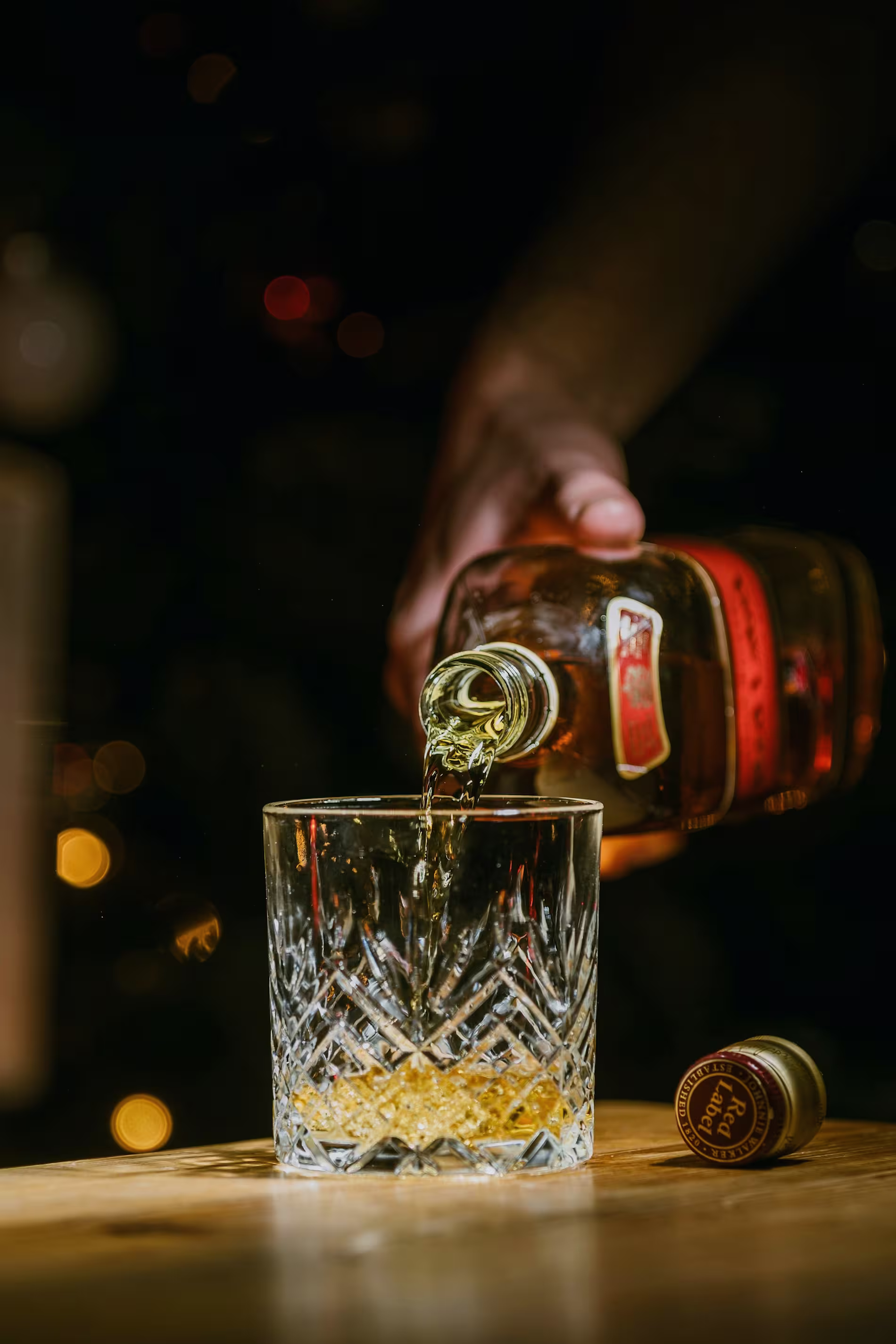 A hand pours whiskey from a bottle into a crystal glass on a wooden surface, with a bottle cap lying nearby and a dark background.