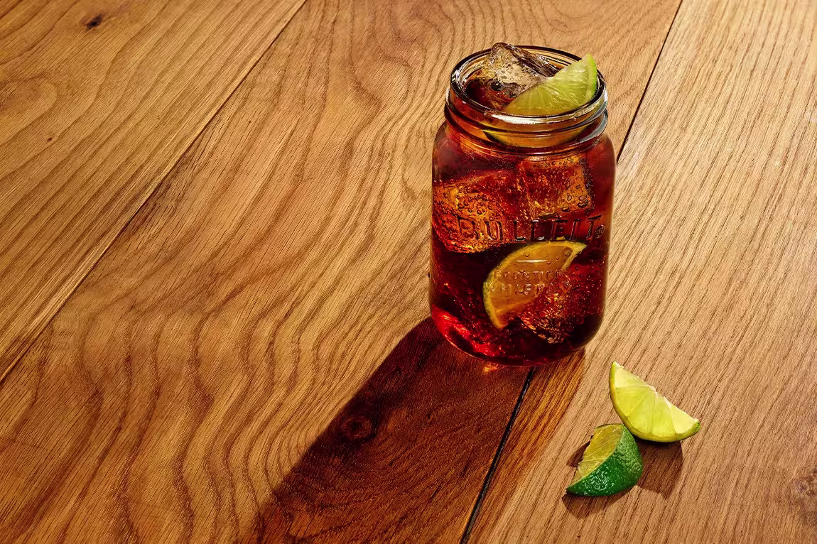 A mason jar filled with a dark iced beverage, lime wedges inside and on top, sits on a wooden surface with extra lime wedges nearby.