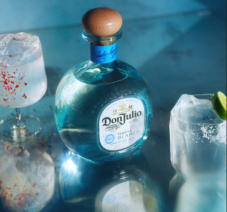 En la imagen se ve la botella de Don Julio Blanco sobre un fondo celeste. Lo rodean dos vasos con bebidas blancas y decoración.