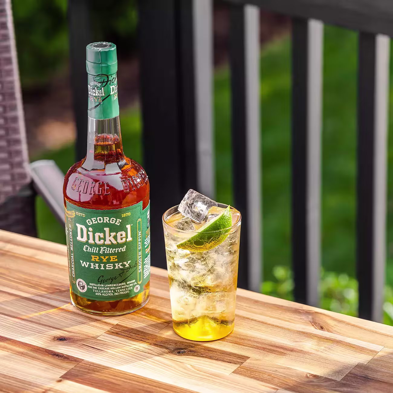 A bottle of George Dickel Rye Whisky and a glass of whisky with ice and a lime wedge sit on a wooden outdoor table.