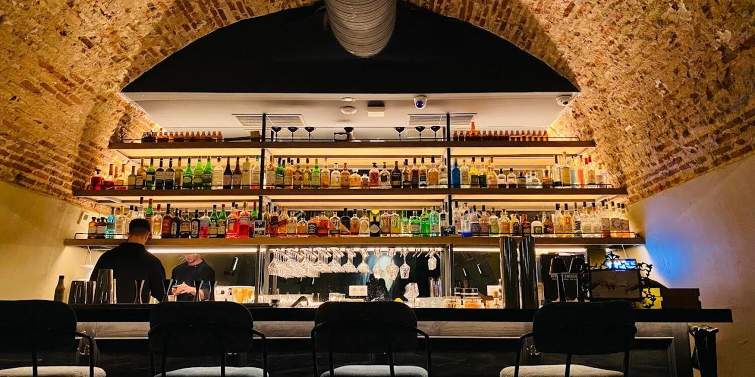El interior de la cockteleria Maldita Gioconda  con un ambiente acogedor y cálido. El techo es de ladrillo arqueado, lo que añade un encanto rústico al lugar. La pared del fondo está llena de estantes con varias botellas de alcohol.
