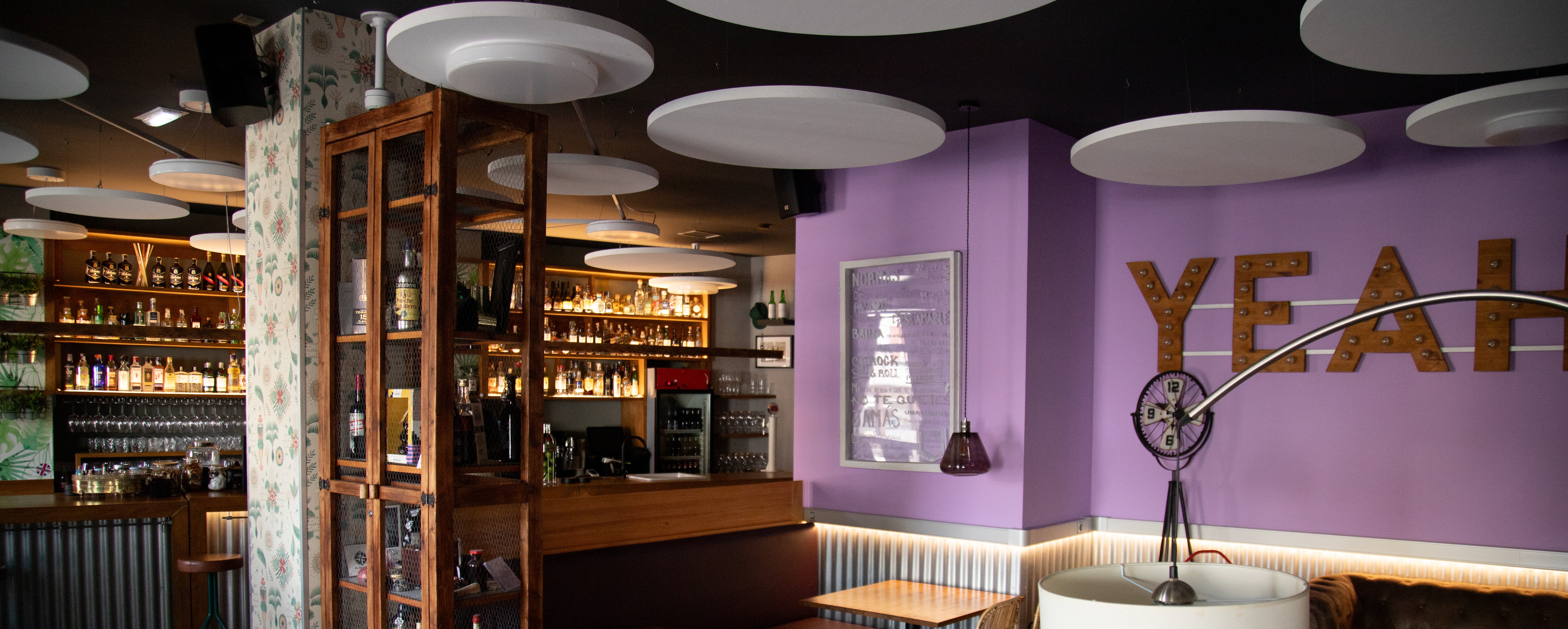 La imagen muestra el interior de Patio de Butacas, con mesas y sillas de madera, un sofá con cojines, luces circulares y un gabinete con botellas. En la pared, una gran pieza decorativa dice “YEAH”. El espacio tiene paredes púrpuras.