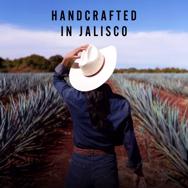 A woman in a wide-brimmed hat walks through an agave field under a clear blue sky. The text "Handcrafted in Jalisco" is displayed above, emphasizing the tequila's origin and artisanal production process.