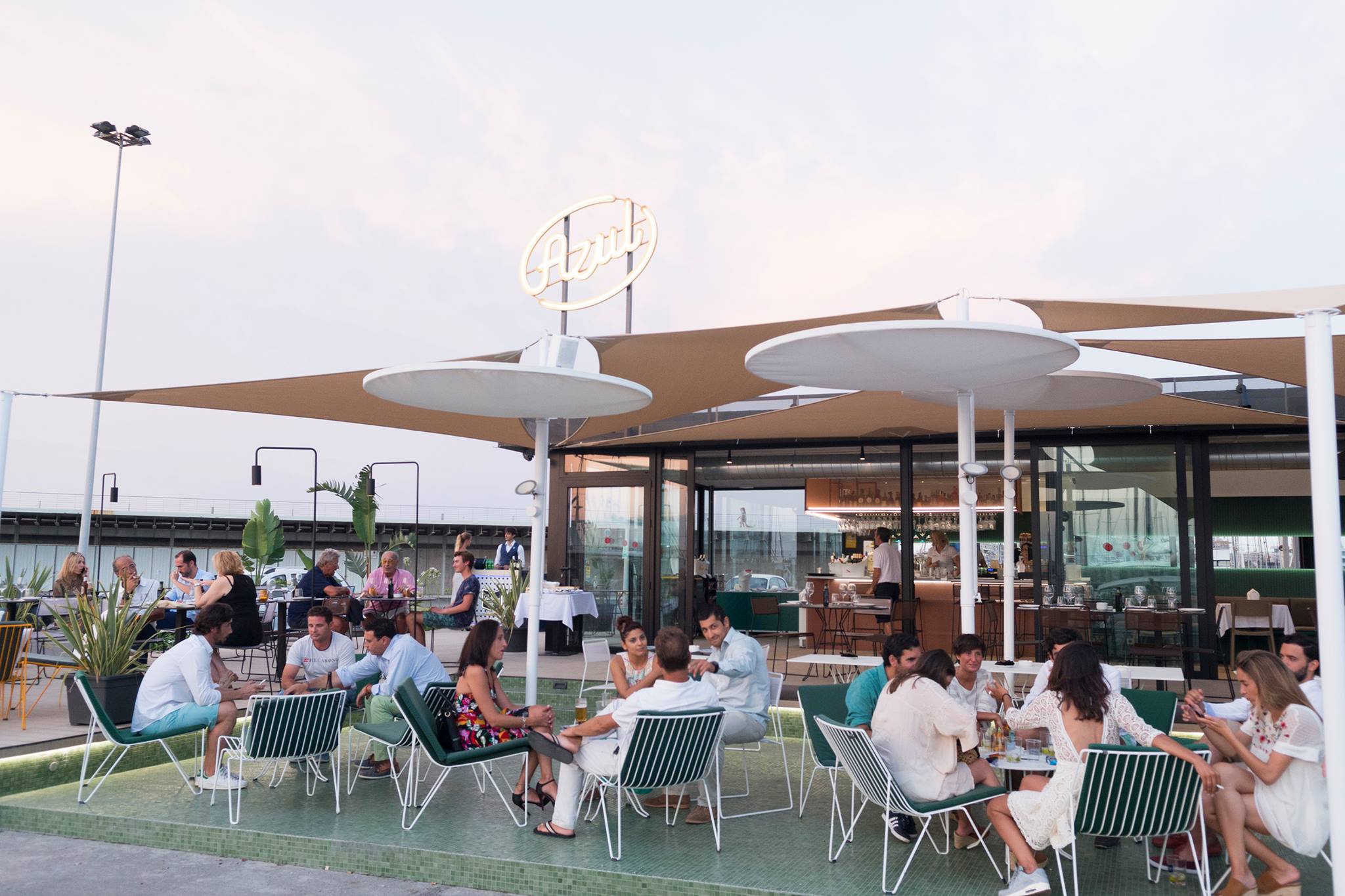 Terraza de Azul Sunset Point al atardecer, con grupos charlando en sillones modernos, sombrillas redondas y un bar acristalado con ambiente relajado junto al mar.