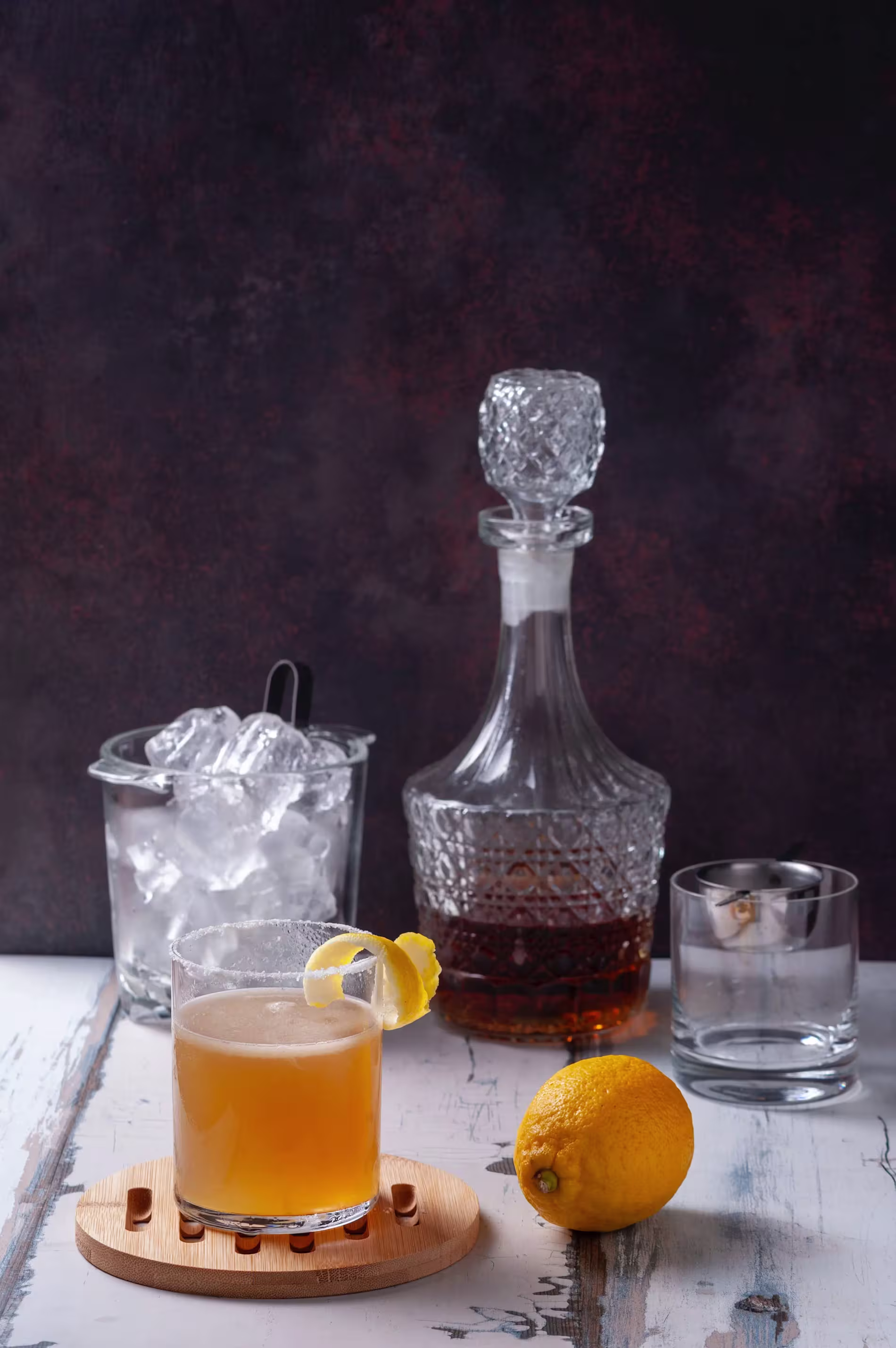 A glass of cocktail garnished with lemon sits on a wooden coaster, with a decanter of liquor, an ice bucket, an empty glass, and a lemon on a table.