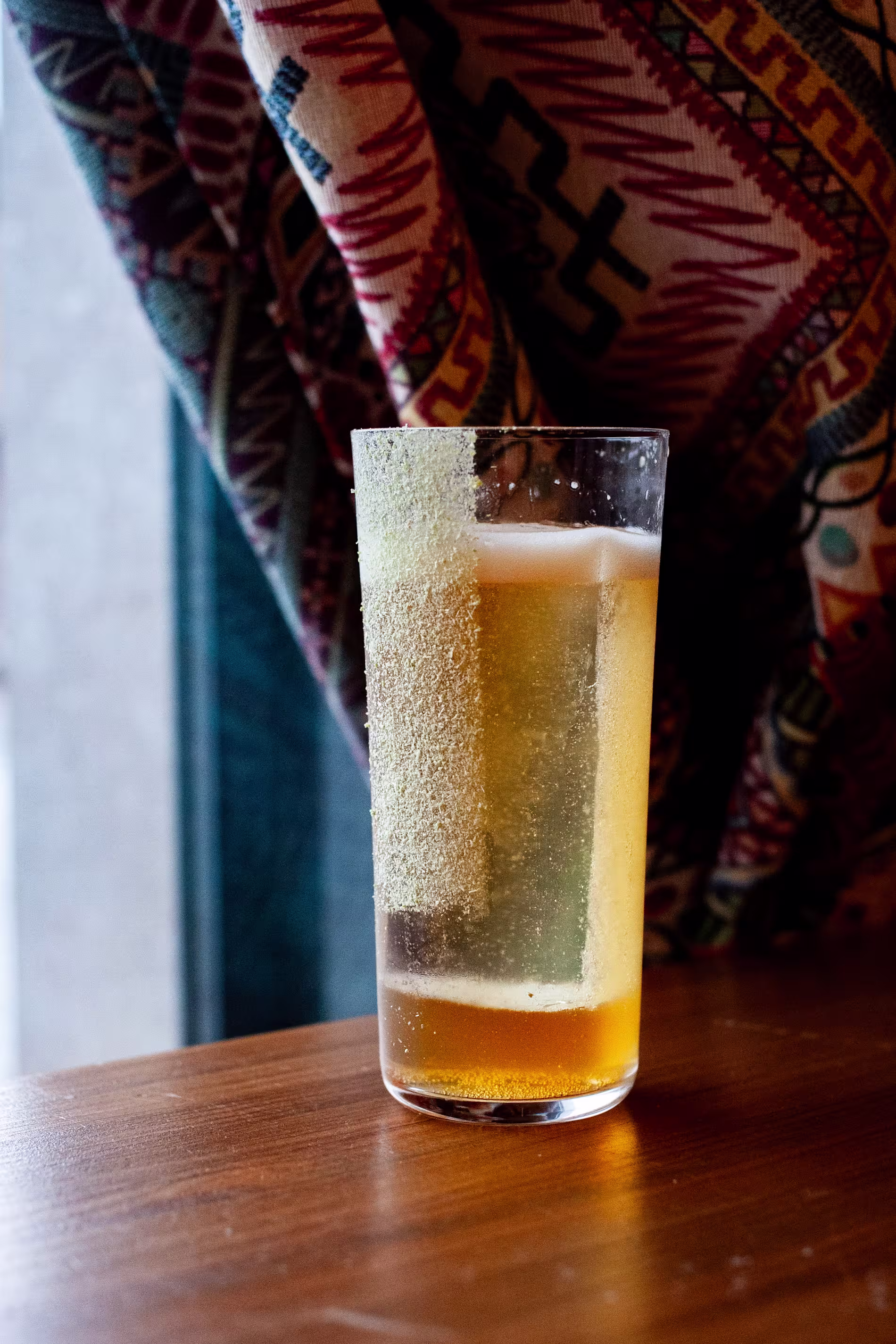 La imagen muestra un primer plano de un vaso transparente de cóctel, posiblemente cerveza. El vaso está colocado sobre una superficie con un fondo desenfocado que es parte de una tela con patrones.