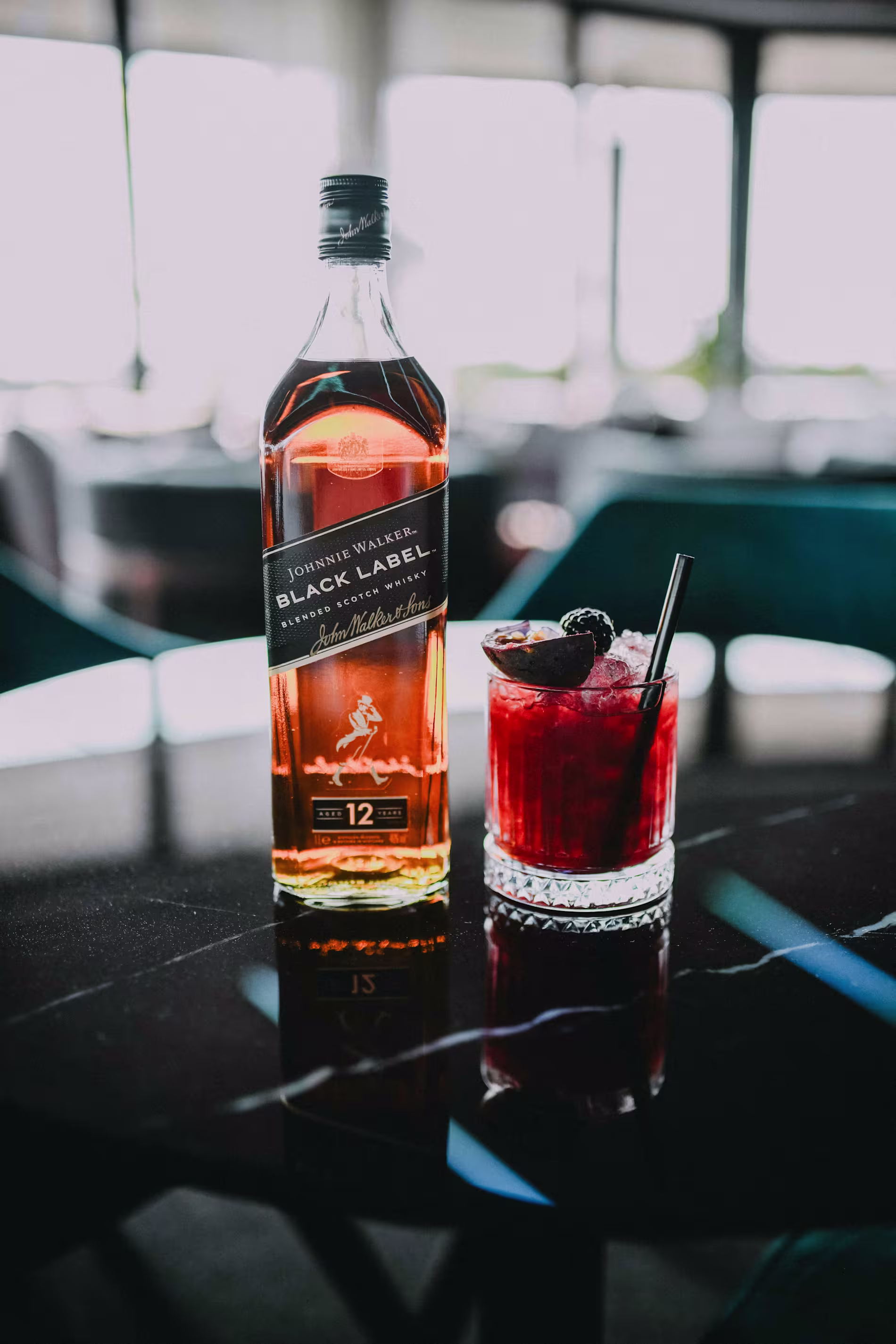 A bottle of Johnnie Walker Black Label whisky stands next to a red cocktail with ice, fruit garnish, and a black straw on a dark reflective table.