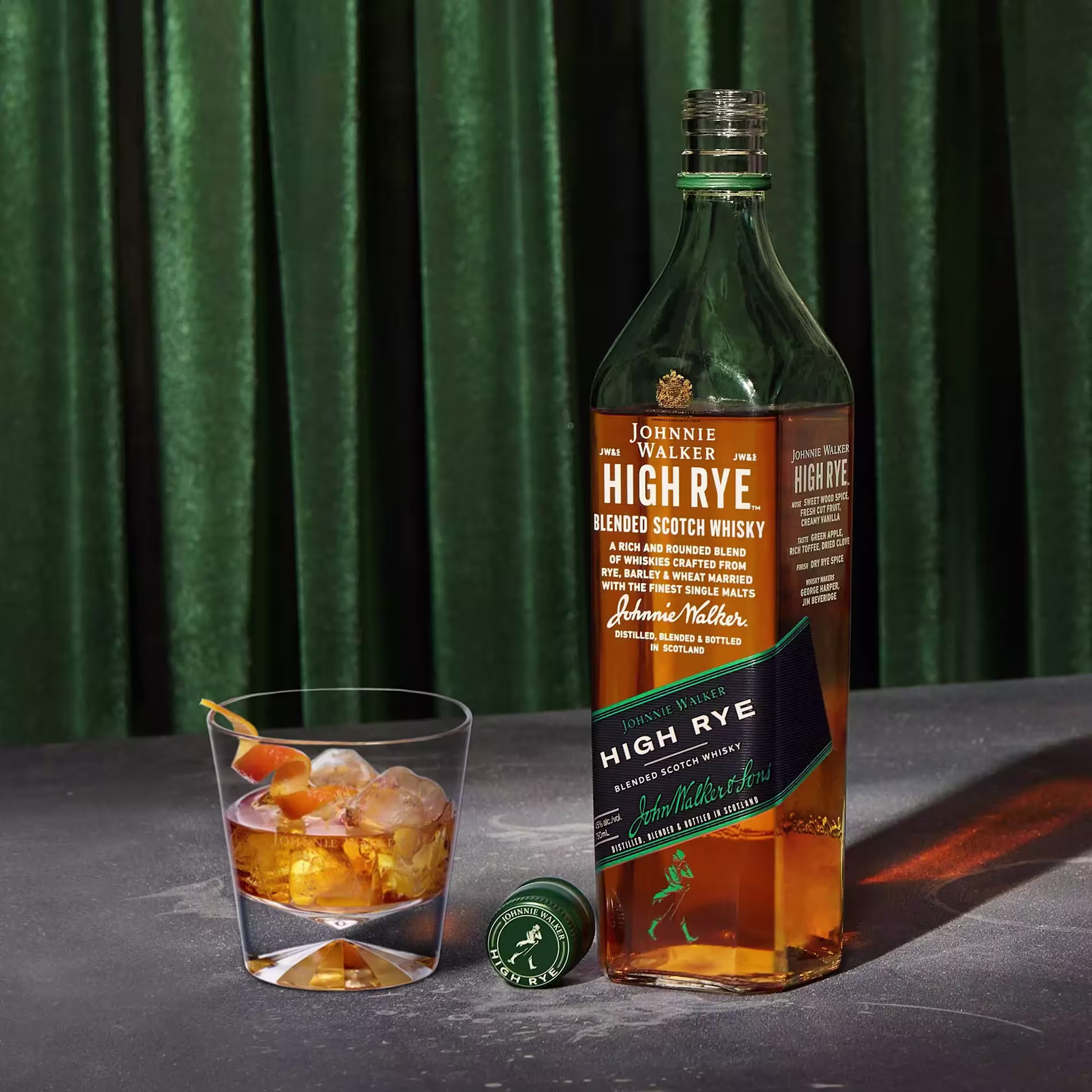 A bottle of Johnnie Walker High Rye Scotch Whisky stands next to a glass of whisky with ice and an orange twist, with a green cap on the table and a green curtain in the background.
