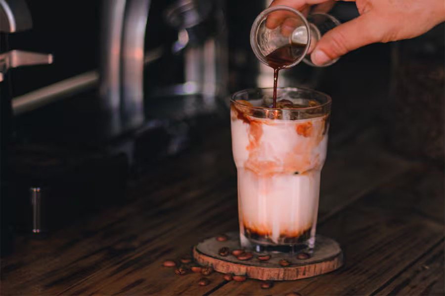 A hand pours espresso into a tall glass of iced milk on a wooden table, with coffee beans scattered nearby.