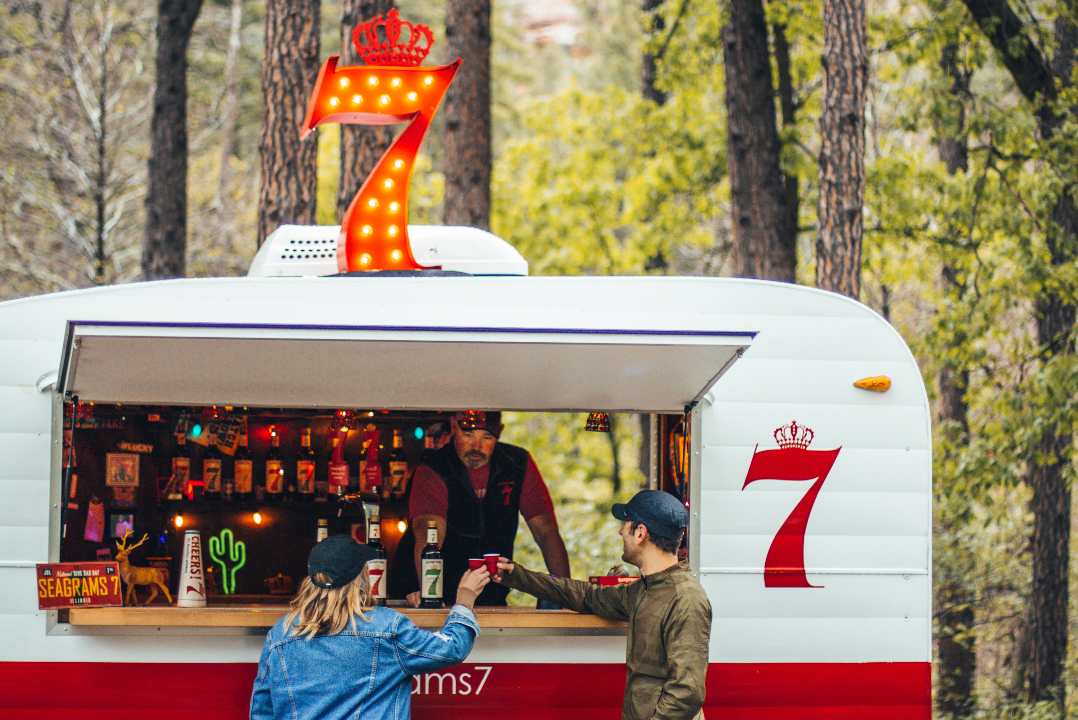 Two people receive drinks from a server at a white and red beverage trailer with a large illuminated number 7 sign and bottles displayed inside, set in a wooded area.