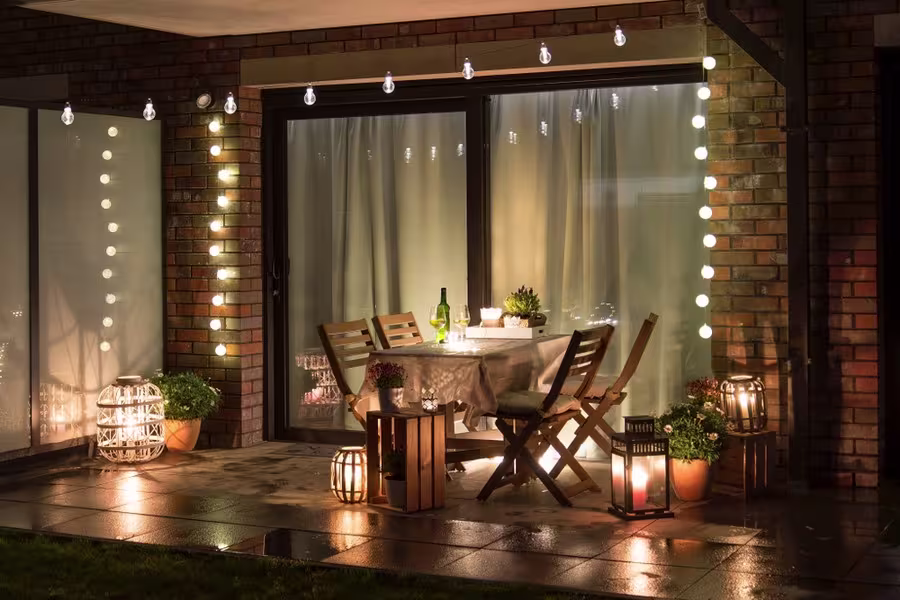 A small patio with a wooden table and two chairs, set for dinner, surrounded by string lights, lanterns, potted plants, and a bottle of wine on the table.