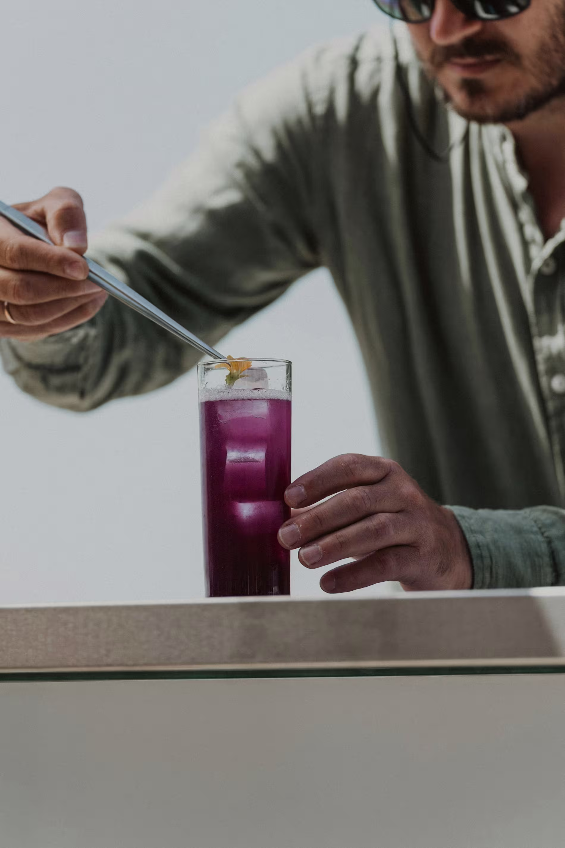 A person in sunglasses garnishes a tall glass of purple drink with ice using tongs, on a light-colored surface.