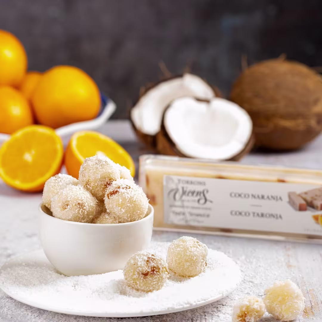 Bolas de coco cubiertas de ralladura fina servidas en un cuenco blanco, acompañadas de naranjas frescas y coco natural, junto a una barra de turrón de coco y naranja de Torrons Vicens.