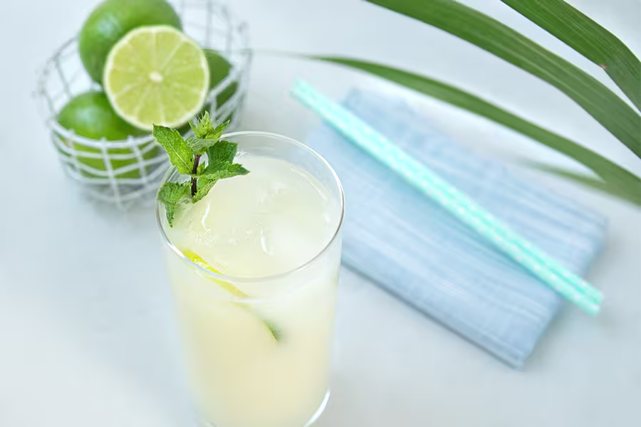 A glass of iced lime drink garnished with mint sits on a table beside whole limes in a wire basket, two blue straws, and a blue napkin.