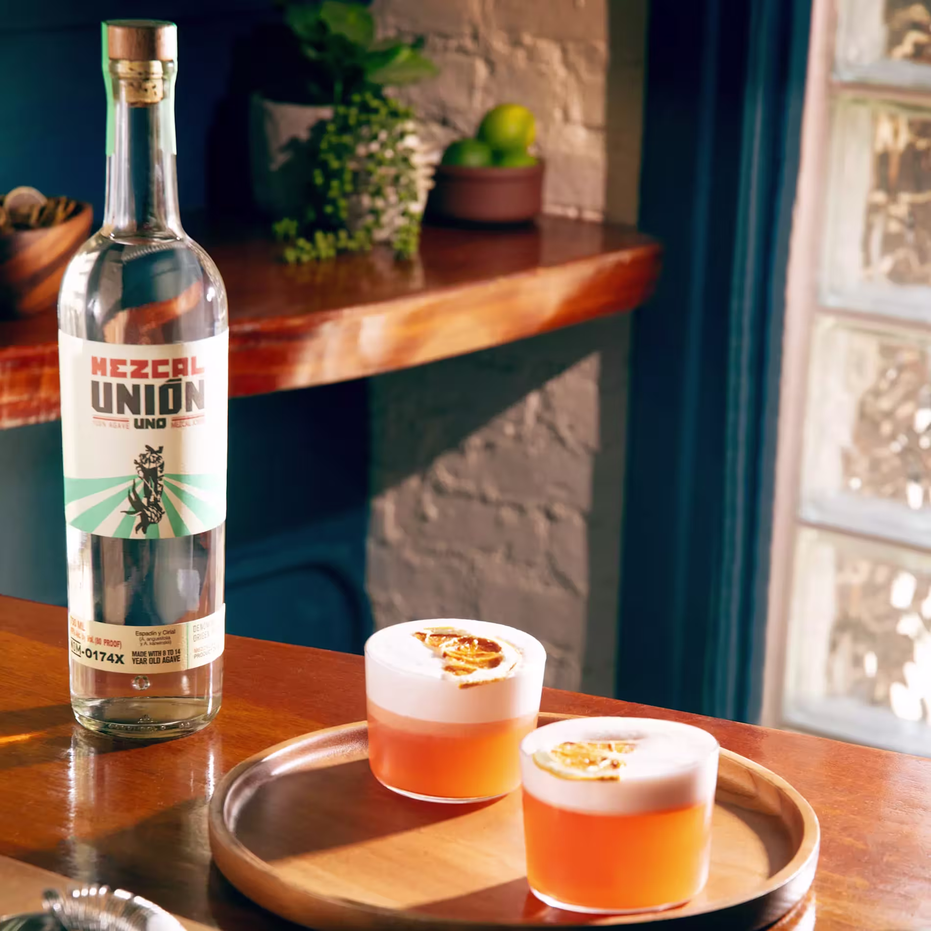 A bottle of Mezcal Unión sits next to two cocktails with frothy tops and dried citrus garnishes on a wooden tray in a warmly lit room.