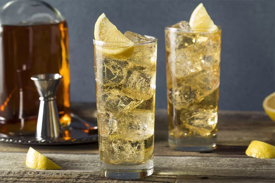 Two tall glasses filled with an amber drink, ice cubes, and lemon wedges on a wooden surface, with a liquor bottle and jigger in the background.