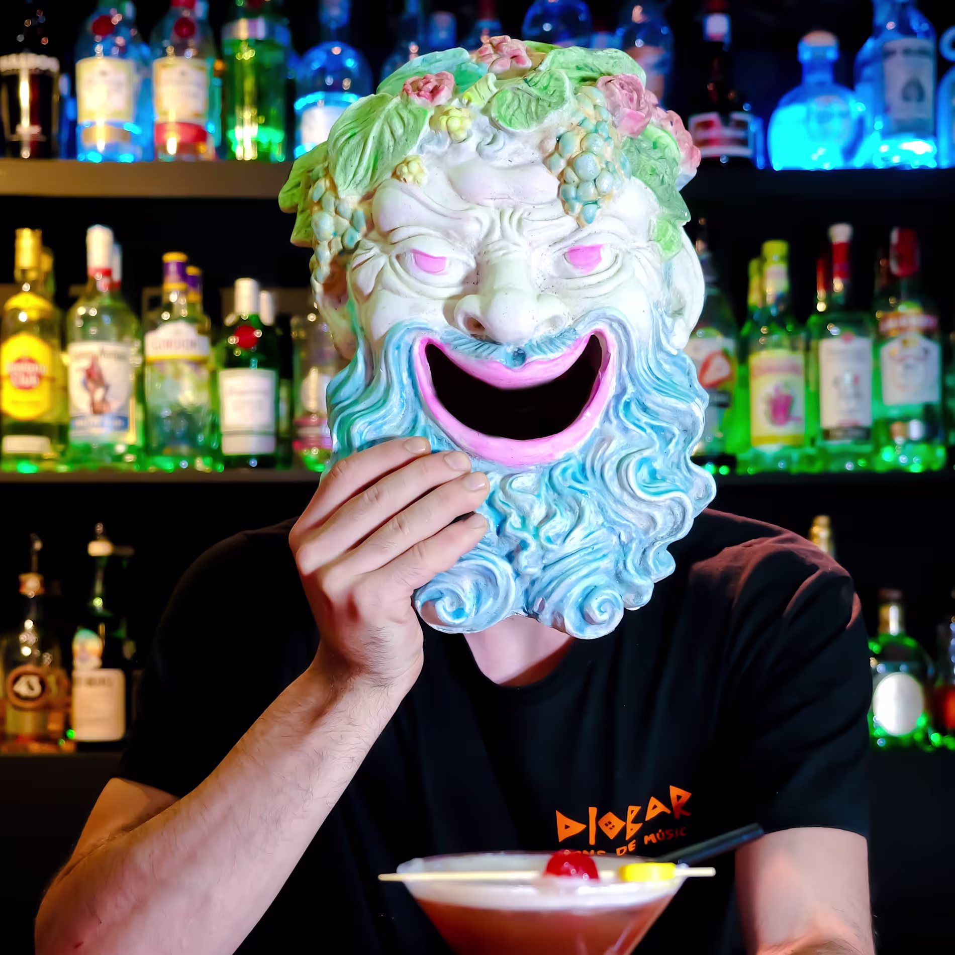 La imagen muestra a una persona con una camiseta negra con la palabra “DIOBAR” visible, de pie detrás de una barra con varias botellas de alcohol en el fondo. La persona lleva una máscara de teatro griego. Delante de esta persona hay un vaso de cóctel.