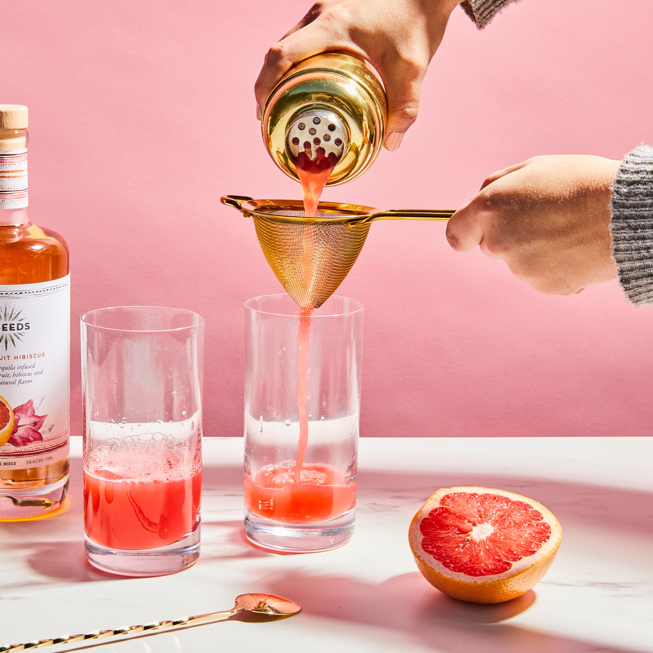 A person strains grapefruit juice through a gold sieve into a tall glass. 
