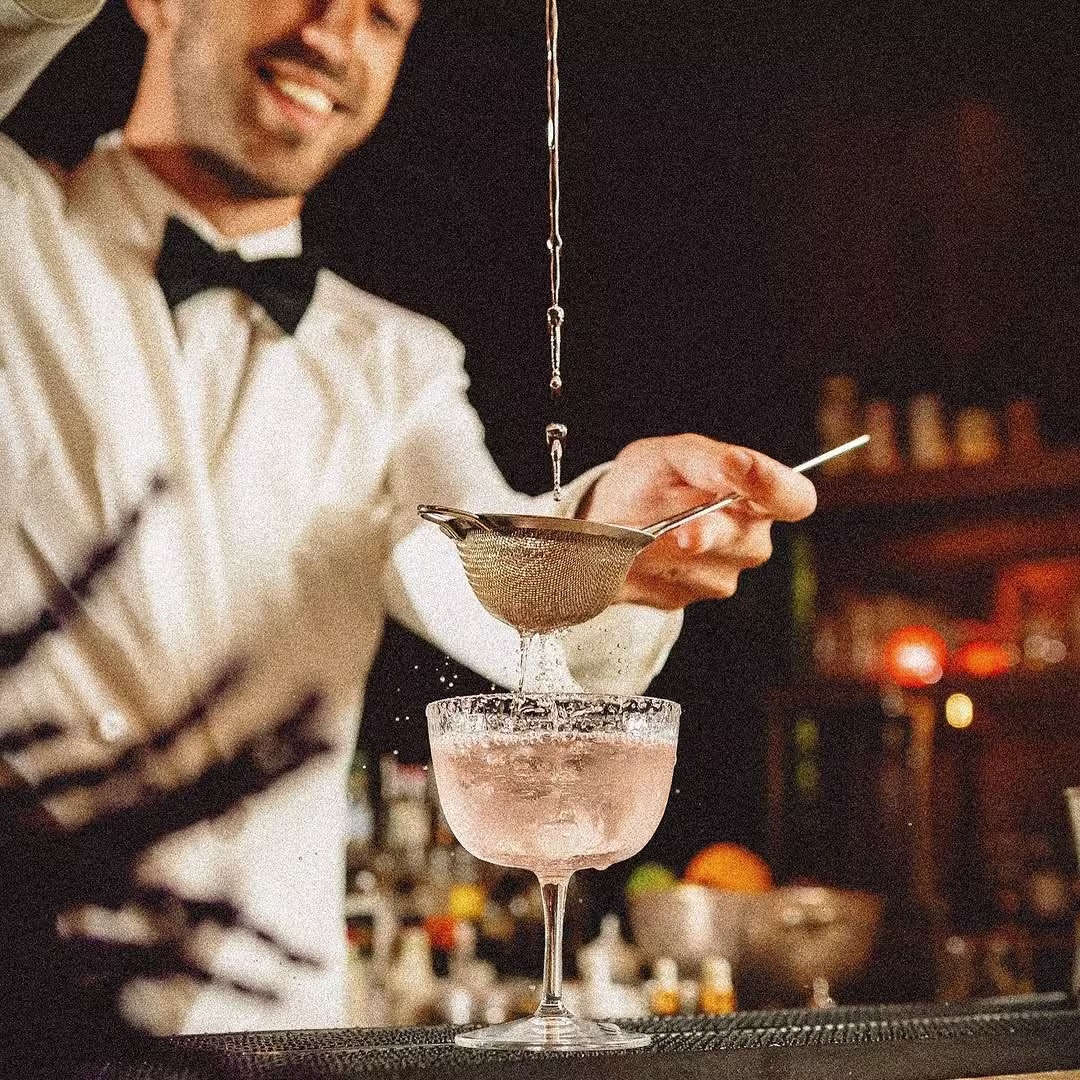 La imagen muestra a un bartender sirviendo un cóctel en una copa elegante con una fina malla para colar, mientras se observa un fondo de botellas y luces cálidas en un bar sofisticado.