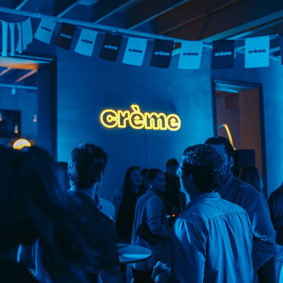 En la imagen se ve el logo de Creme, en luces neon, con algo de gente a su alrededor. El contexto esta en tonos azules.