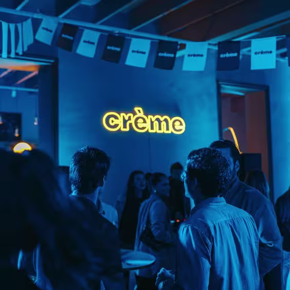 En la imagen se ve el logo de Creme, en luces neon, con algo de gente a su alrededor. El contexto esta en tonos azules.
