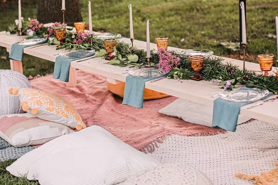 A low picnic table set with glass plates, blue napkins, amber glasses, candles, and floral greenery, surrounded by pillows on blankets outdoors.
