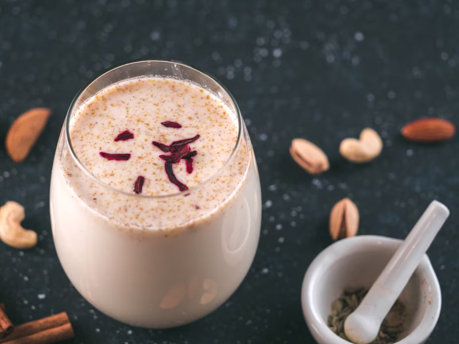 A glass of creamy beverage topped with saffron strands, surrounded by scattered almonds, cashews, and a small bowl with a pestle.