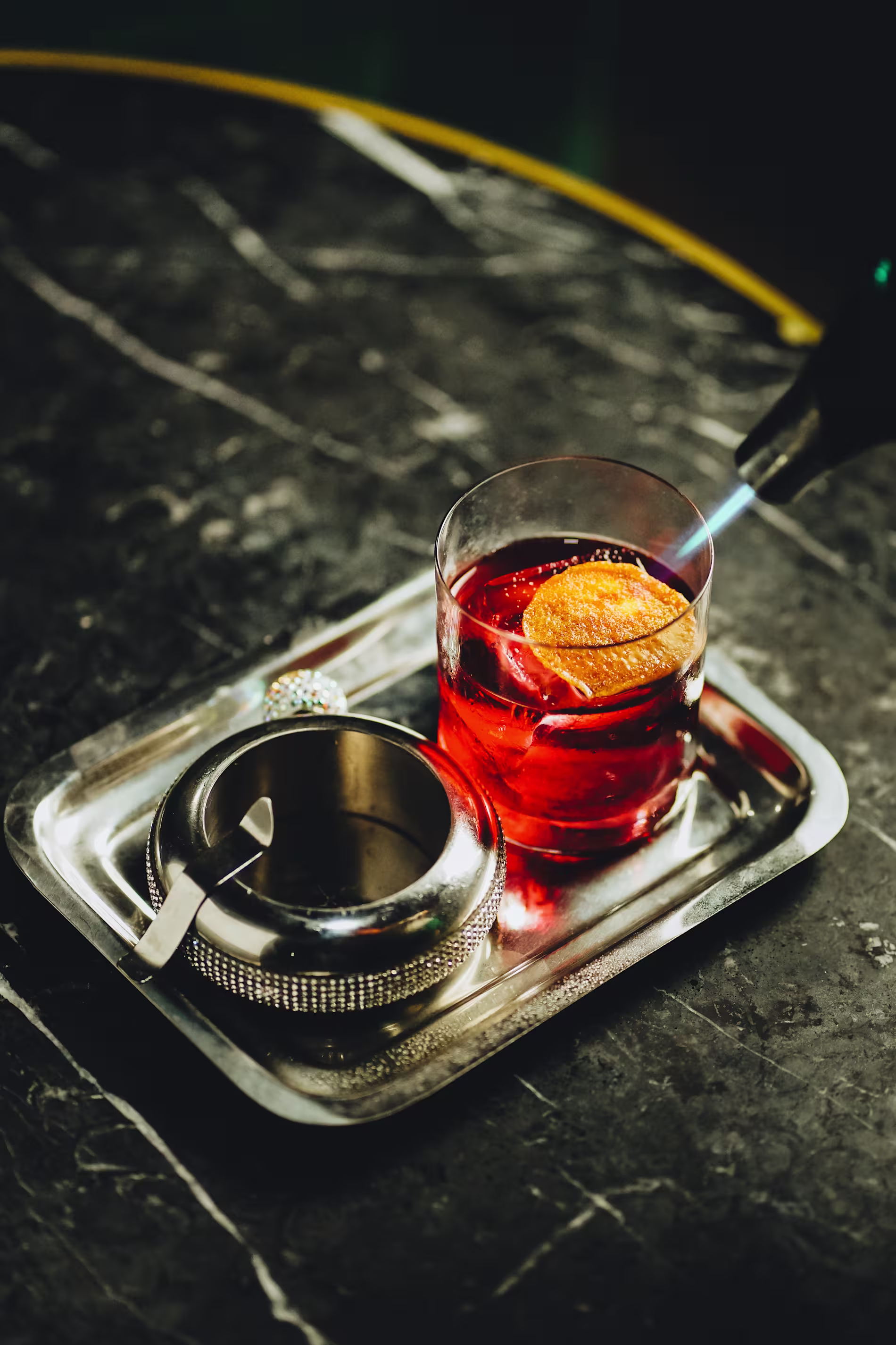 La imagen muestra un vaso con un cóctel, con una rodaja de cítrico en el borde y un agitador de metal dentro. El vaso está colocado sobre una pequeña bandeja rectangular junto a un cecinero.