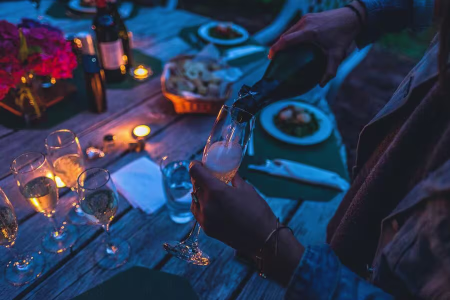 A person pours champagne into a glass at an outdoor dinner table set with food, candles, and flowers in the evening.