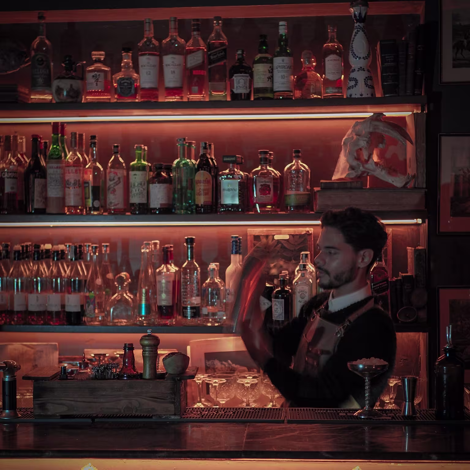 La imagen muestra el interior de Jacks Library con varias botellas de alcohol exhibidas en estantes. Hay una persona de pie detrás del mostrador de la barra agitando un cóctel. La iluminación es tenue y crea una atmósfera íntima
