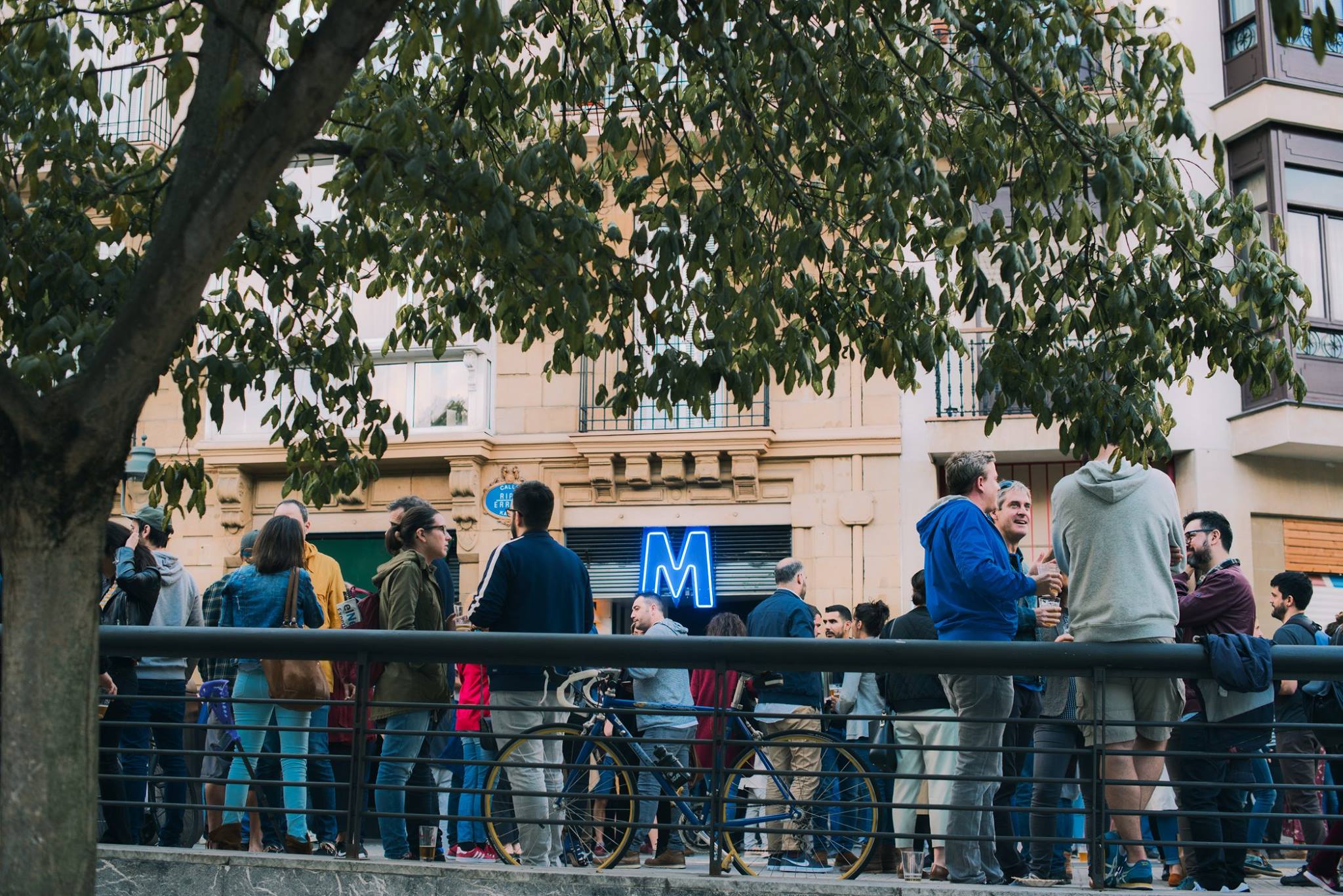 Exterior de Muelle con grupos de personas charlando y bebiendo junto a una barandilla, bicicletas apoyadas y un cartel azul con la letra M al fondo.