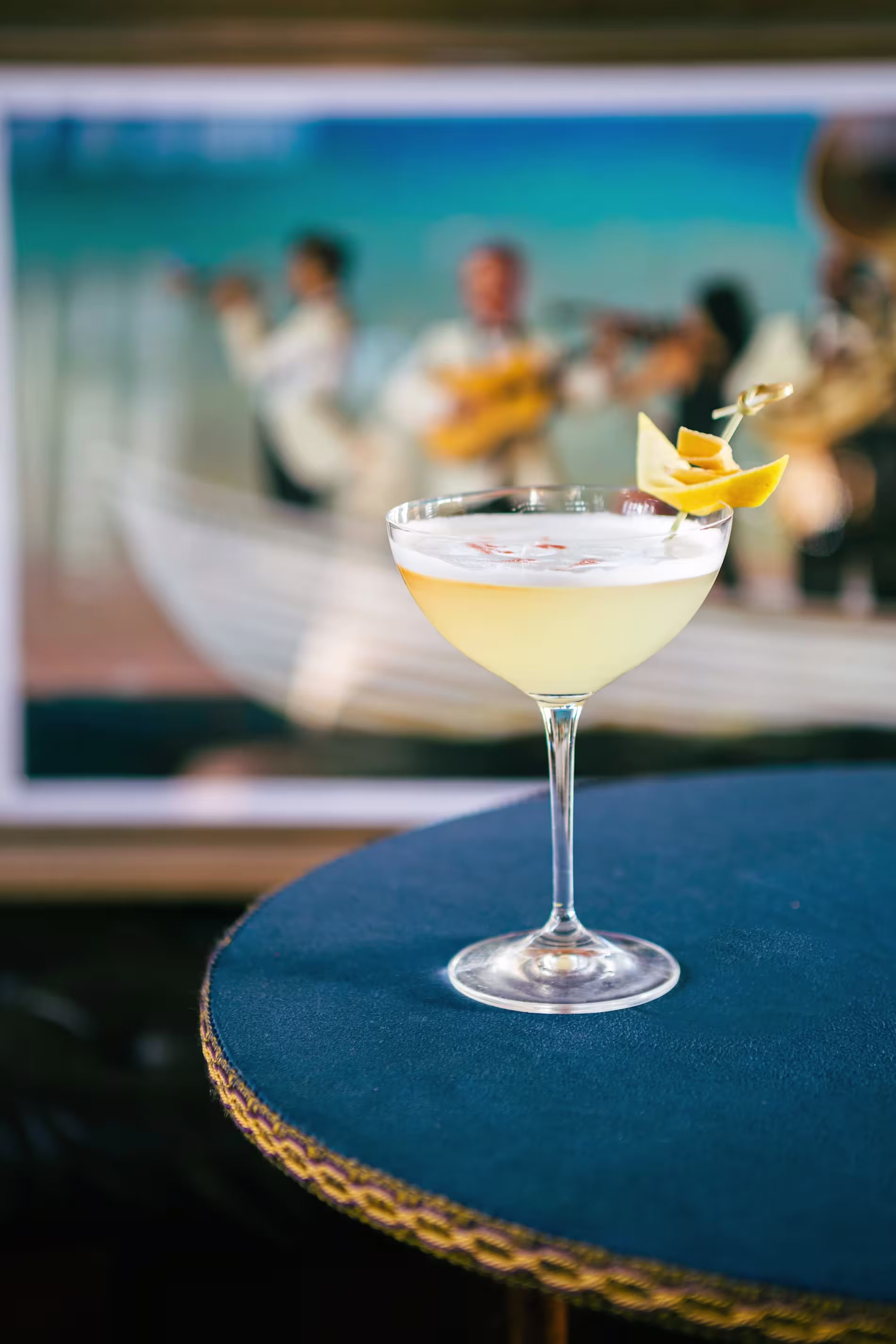 A pale yellow cocktail garnished with a citrus twist sits on a blue table, with a blurred image of musicians in the background.
