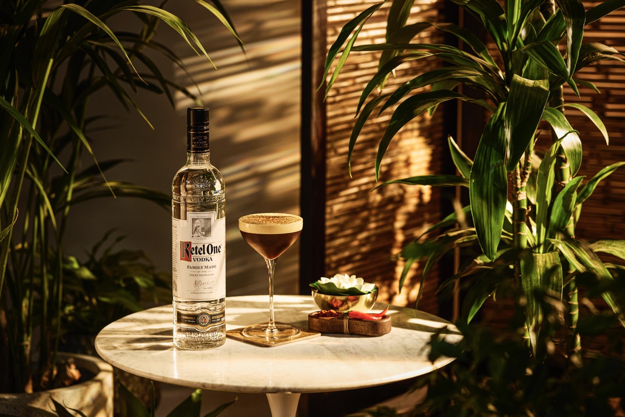 A bottle of Ketel One vodka and a cocktail glass sit on a marble table, surrounded by green plants and sunlight, with a small dish and flower nearby.