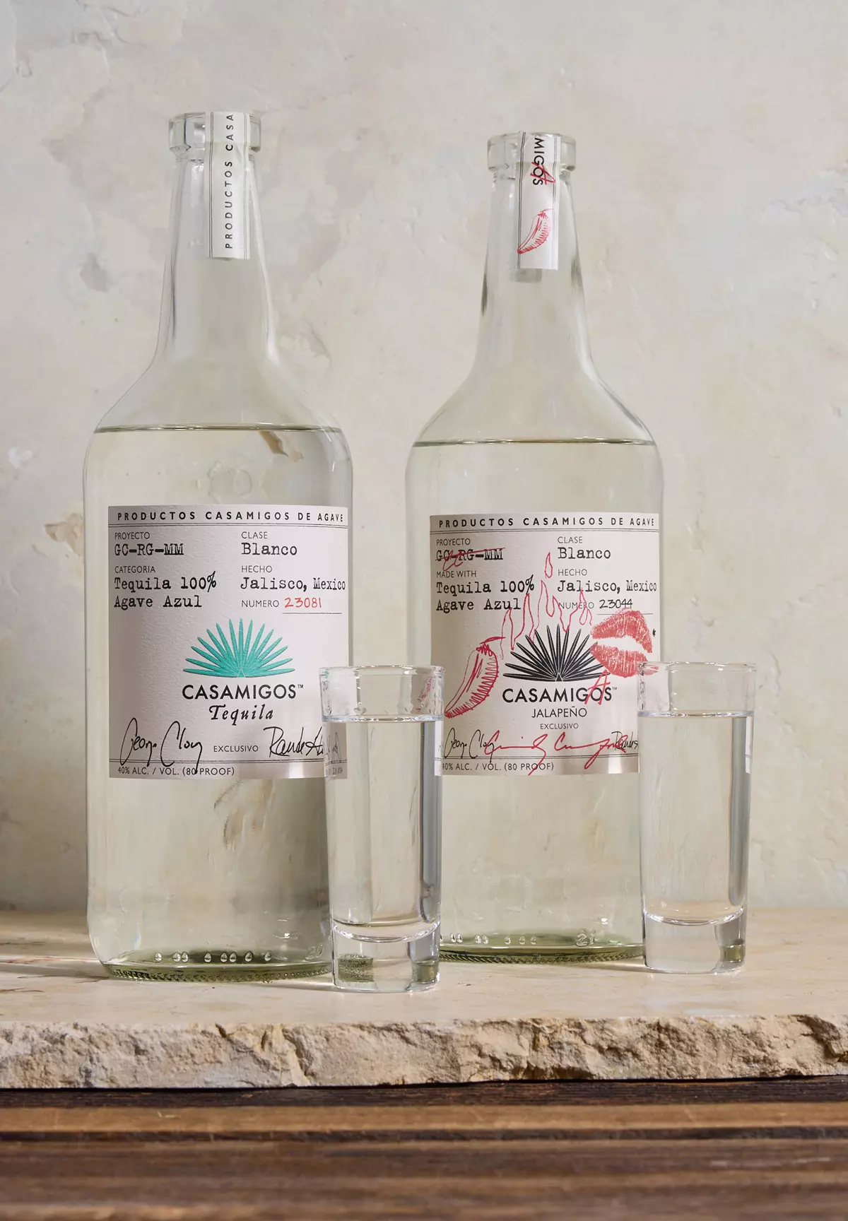 Two bottles of Casamigos tequila, one Blanco and one Jalapeño flavor, stand side by side with a filled shot glass in front of each, on a stone surface against a neutral background.