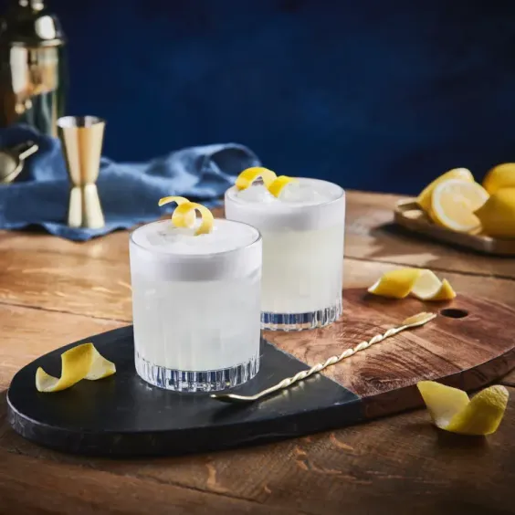 Dos vasos de Gin Sour sobre una tabla, con un batidor y cáscaras de limón