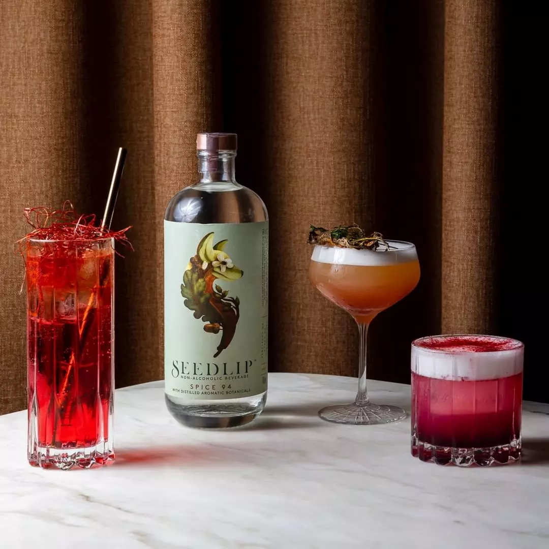 A bottle of Seedlip Spice 94 is displayed on a marble table with three colorful cocktails in different glasses, set against a brown curtain background.
