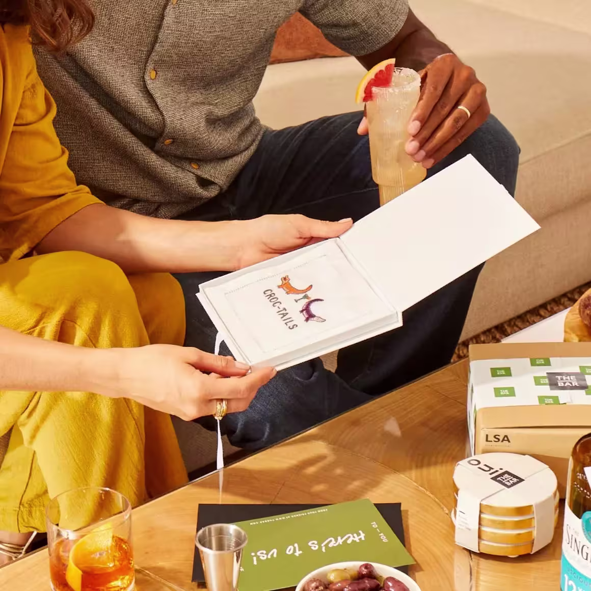 Two people sit on a couch; one holds a drink, the other opens a cocktail recipe booklet on a table with drinks, a booklet, and cocktail-making items.