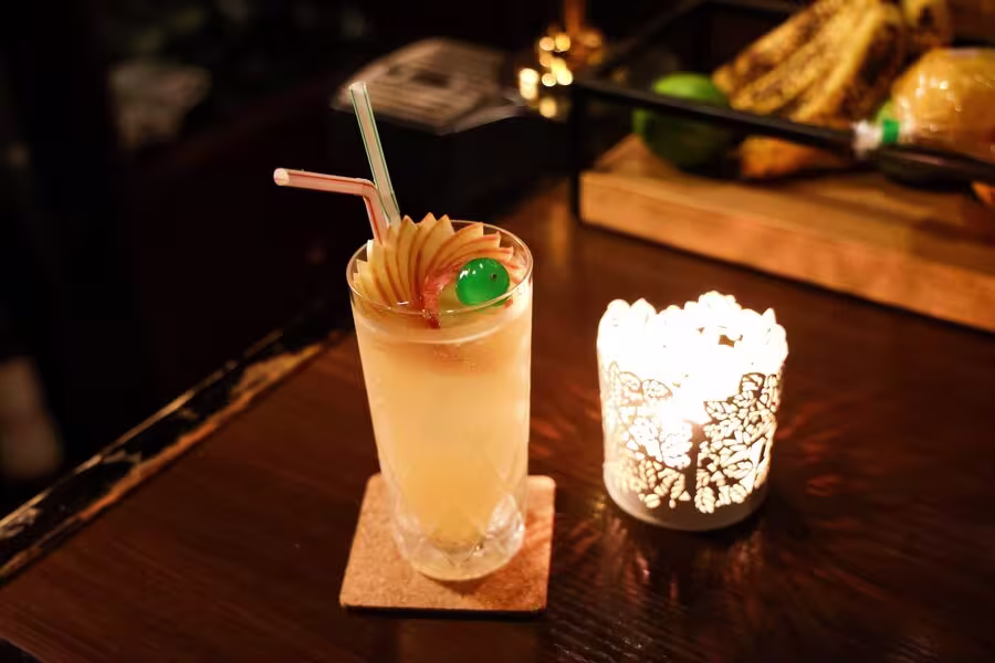 A cocktail garnished with an apple fan and a green cherry sits on a coaster next to a lit decorative candle on a dark wooden table.